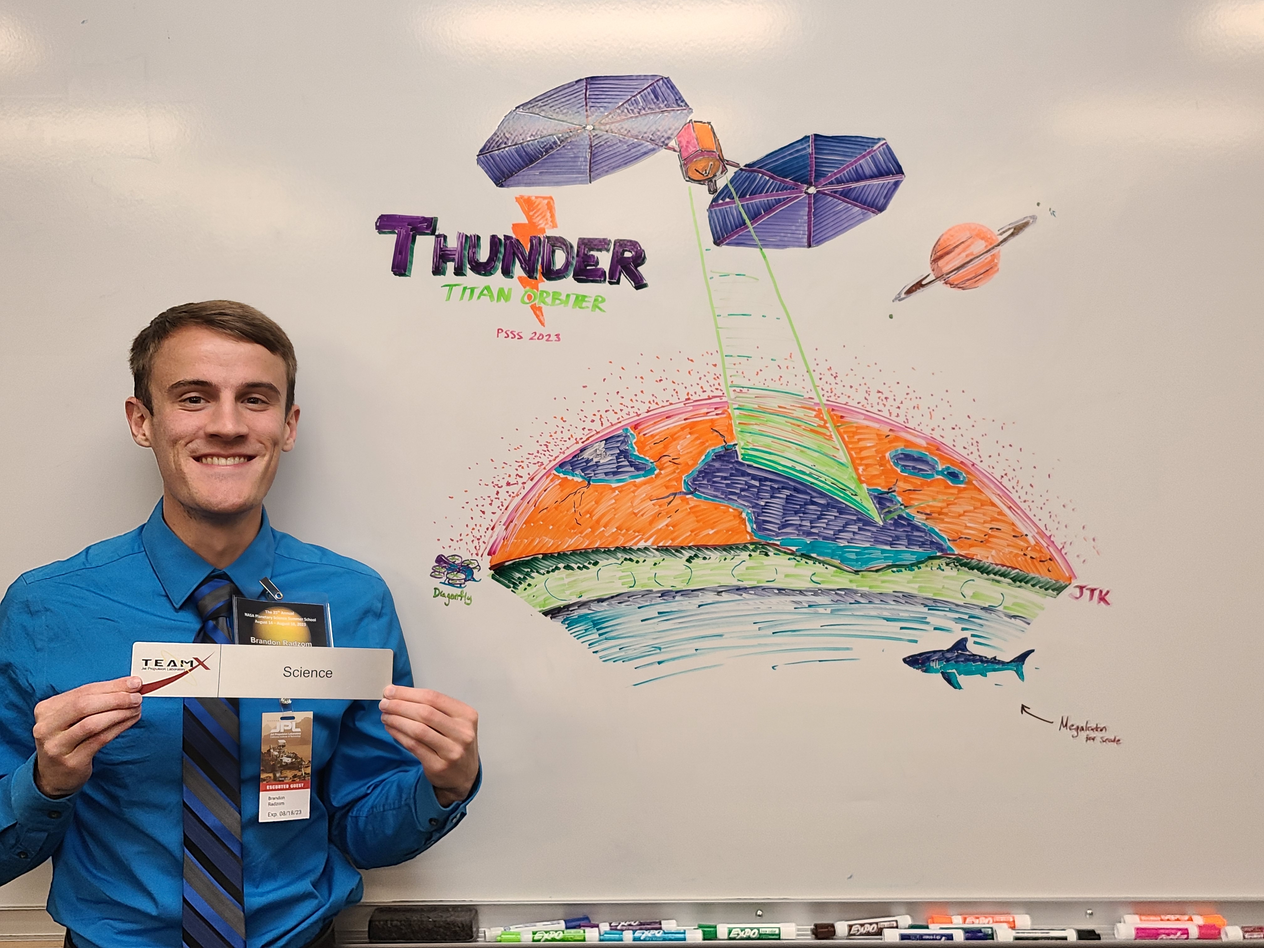 Picture of me holding my subsytem role plack in front of a whiteboard sketch of our mission concept, THUNDER, at JPL.